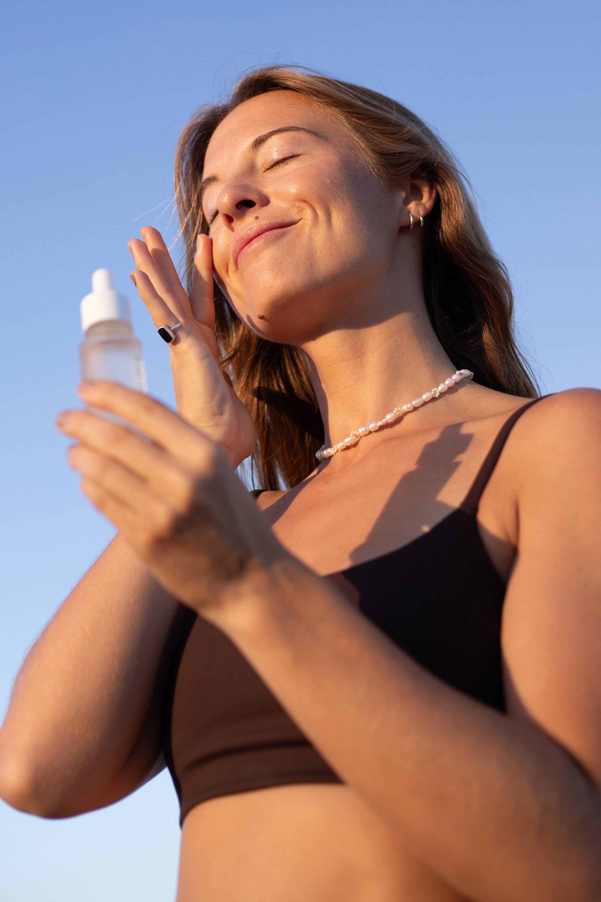 Woman applying burnd restore hydrating skin serum on a clear blue sky background