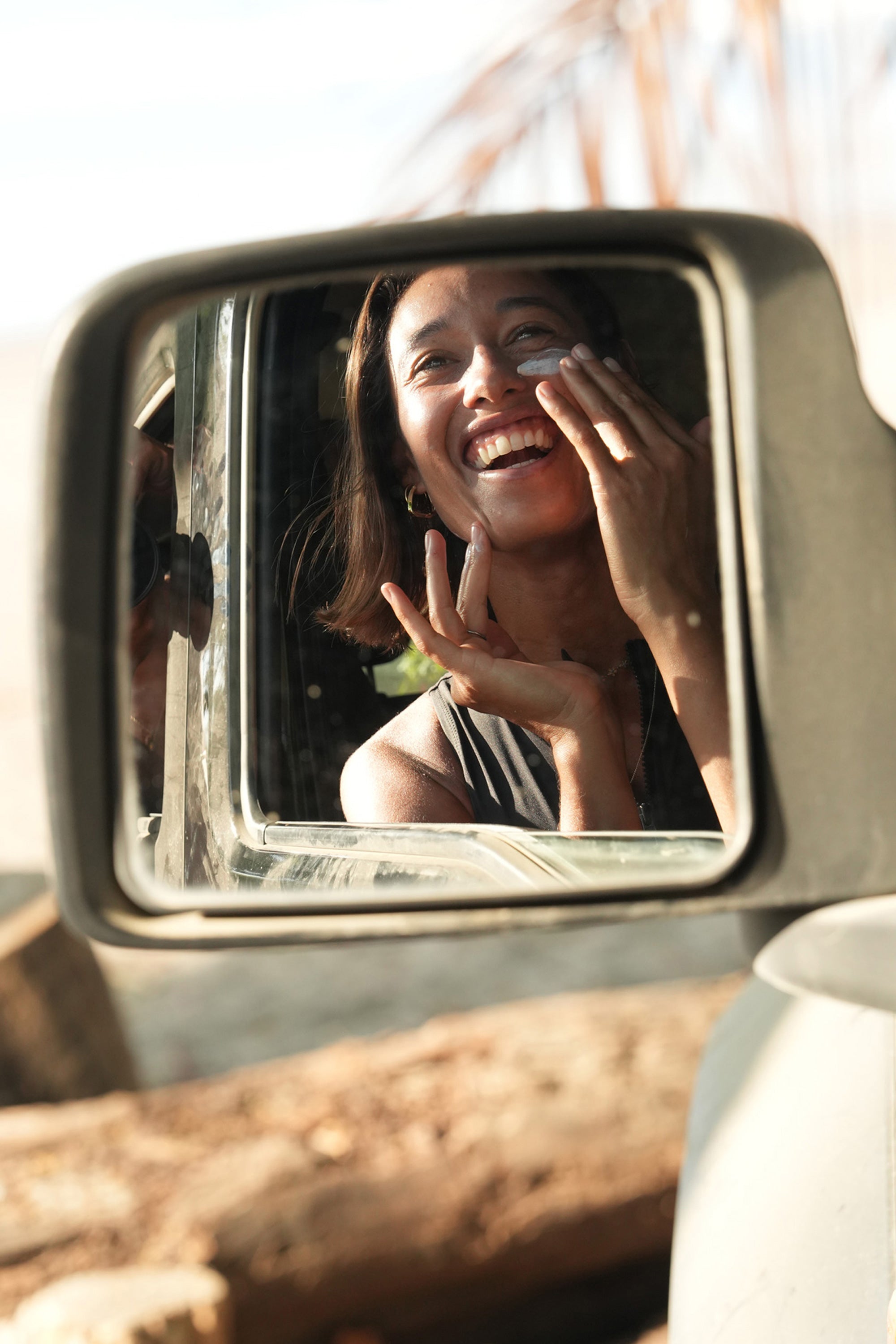Woman applying burnd high performance sunscreen in a car's side mirror with a natural outdoor background
