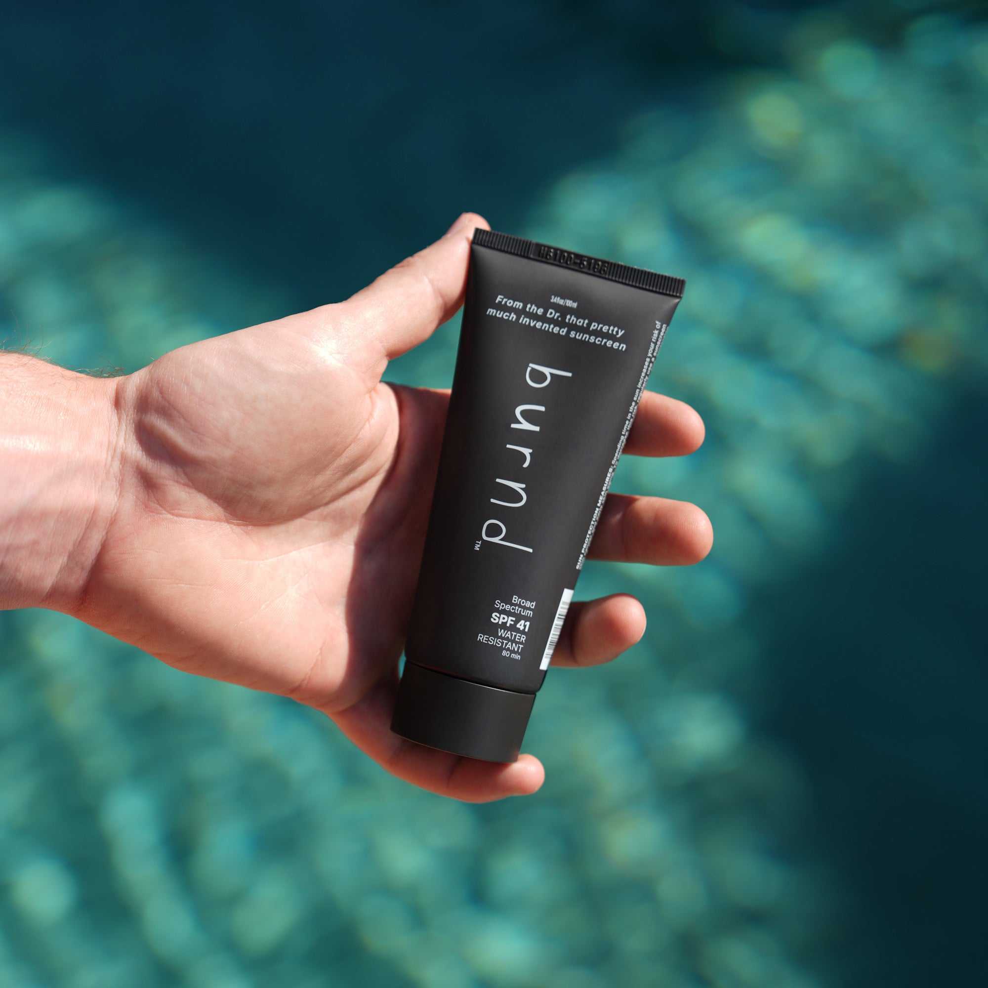 Hand holding a tube of 'burnd SPF41' sunscreen with a blurred blue water background