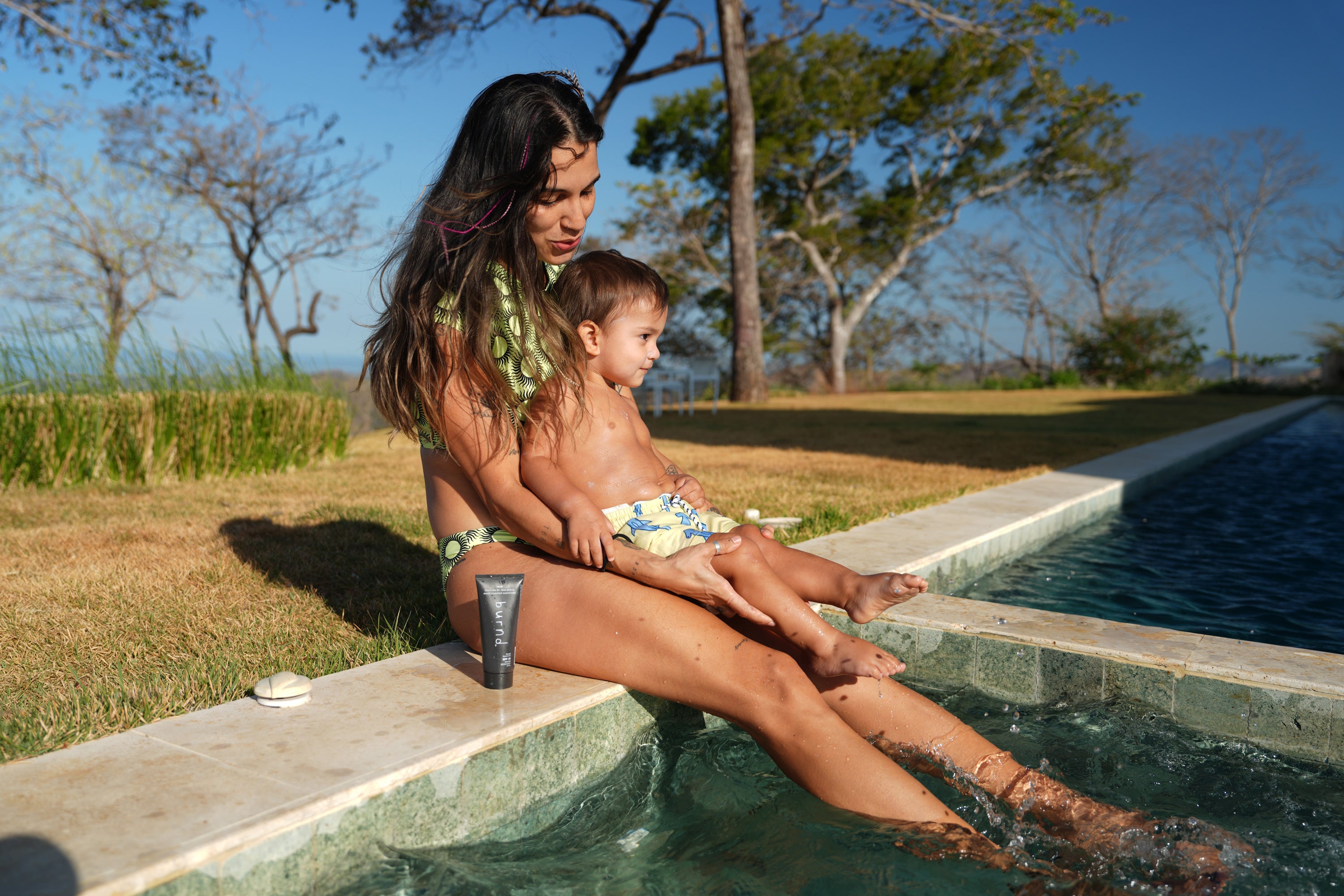 Woman and child sitting by a pool with trees in the background, next to a tube of burnd sunscreen, that is tailored for kids due to the ease of application